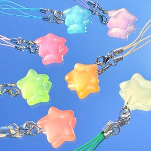 May include: A collection of colorful star-shaped charms in shades of pink, orange, green, yellow, and blue. Each charm is attached to a silver clasp and a matching colored cord. The charms are displayed against a reflective blue surface.