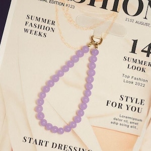 Puede incluir: Un dije de teléfono con cuentas de color lavanda con un cierre dorado. El dije está hecho de pequeñas cuentas redondas y está unido a una pestaña de plástico transparente. El fondo es una página de revista con el texto "Summer Fashion Weeks" y "Style for You."