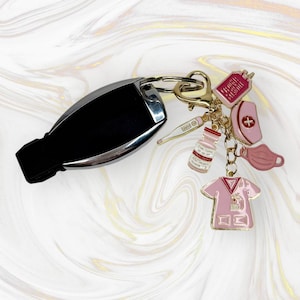 Könnte beinhalten: Ein rosa und goldener Schlüsselanhänger mit verschiedenen Anhängern, darunter ein Stethoskop, eine Pillenflasche, ein Thermometer, eine Gesichtsmaske, eine Krankenschwestermütze und eine Krankenschwesteruniform. Der Schlüsselanhänger ist an einem schwarzen Autoschlüssel befestigt.
