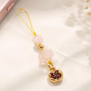 May include: A phone charm with a yellow loop, pink beads, gold detailing, and a gold-coloured pomegranate charm with red accents. The charm is designed to attach to a phone or small device.