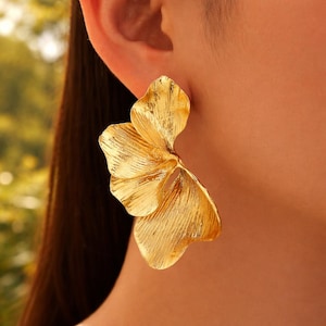 Puede incluir: Primer plano de un pendiente dorado. El pendiente presenta un diseño texturizado y en capas que se asemeja a una flor o una hoja estilizada. El color dorado tiene un acabado cepillado, lo que agrega profundidad y dimensión. Un accesorio llamativo.