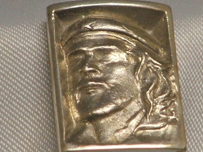 May include: A silver ring with a detailed portrait of a person wearing a hat. The ring is rectangular in shape and has a smooth, polished finish.