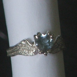 May include: A silver ring with a blue gemstone and two wings on either side of the stone.