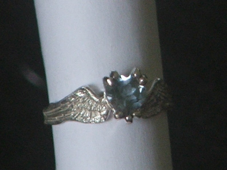May include: A silver ring with a blue gemstone and two wings on either side of the stone.