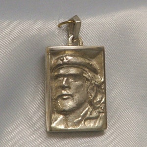 May include: A silver pendant featuring a rectangular portrait of a man with a cap. The pendant has a gold loop at the top for attaching a chain.