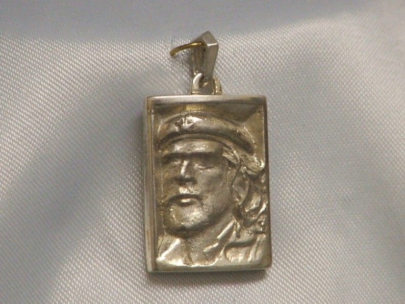 May include: A silver pendant featuring a rectangular portrait of a man with a cap. The pendant has a gold loop at the top for attaching a chain.
