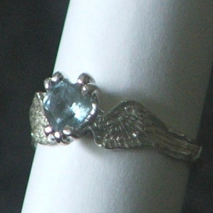 May include: A silver ring with a blue gemstone set in a claw setting. The ring features a wing design on one side.