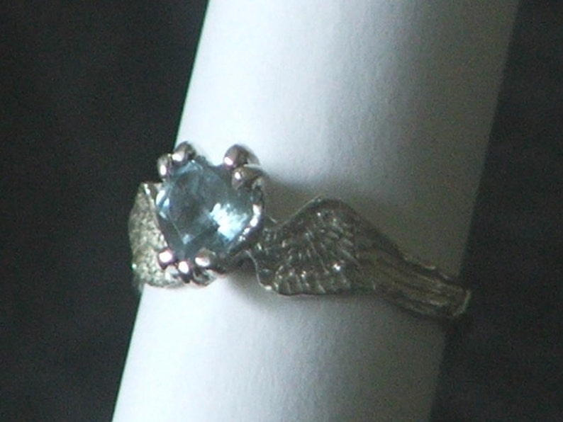 May include: A silver ring with a blue gemstone set in a claw setting. The ring features a wing design on one side.