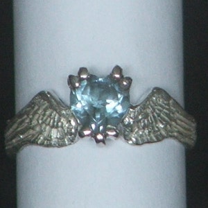 May include: A silver ring with two wings on either side of a heart-shaped blue gemstone.