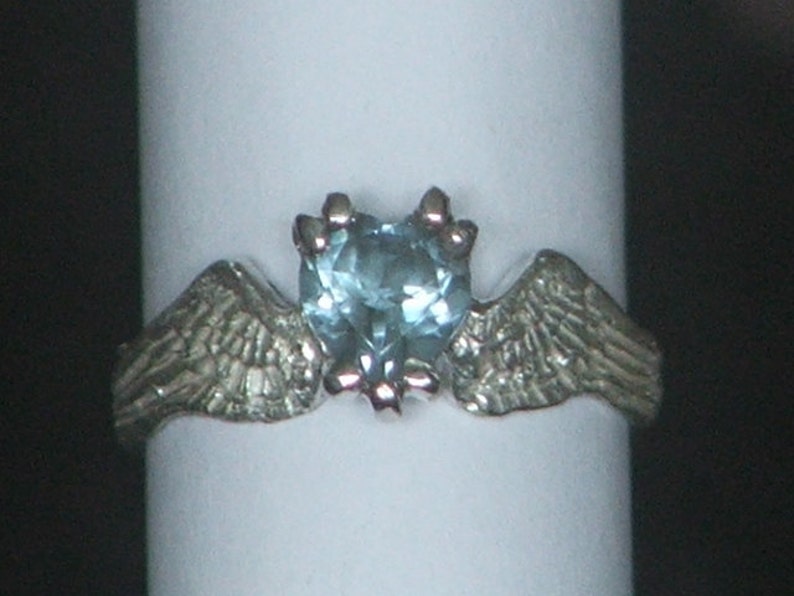 May include: A silver ring with two wings on either side of a heart-shaped blue gemstone.