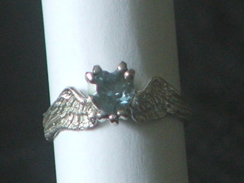 May include: A silver ring with a blue gemstone in the center and two wings on either side.