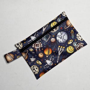 May include: A navy blue zipped pouch with a colourful space-themed pattern. The design includes planets, rockets, astronauts, and robots. The pouch has a black zip and a small fabric loop on the side.