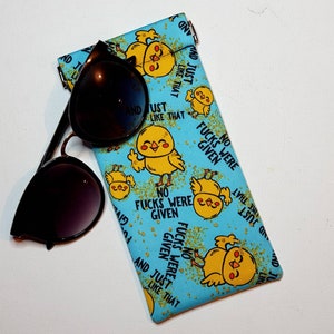 May include: A blue fabric sunglasses case with a repeating pattern of yellow chicks and the text "NO FUCKS WERE GIVEN" and "AND JUST LIKE THAT". Black sunglasses rest on the case.