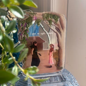 May include: A clear acrylic display featuring a scene with two figures under a tree, with the text "I guess our ripples resonated." The artwork has a painted background with a house and foliage. The base is a decorative, textured design.