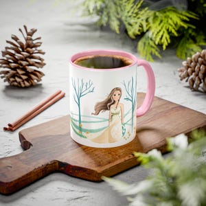 Könnte beinhalten: Eine weiße Keramiktasse mit rosa Innenraum und Henkel, gefüllt mit Kaffee. Die Tasse zeigt eine skurrile Illustration einer Frau in einem fließenden Kleid auf einem Feld mit Bäumen. Die Tasse steht auf einem Holzschneidebrett mit Zimtstangen und Tannenzapfen.