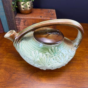 May include: A vintage ceramic teapot with a light green body and a brown handle and lid. The teapot features embossed floral detailing and a curved spout. The handle is arched, and the lid has a small knob. The teapot is a decorative kitchenware item.
