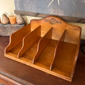 May include: A vintage wooden desk organizer with four angled compartments. The organizer is made of light brown wood and features a curved top with a decorative emblem. The design is ideal for organizing mail, documents, or office supplies.