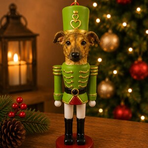 Può includere: Una statuetta schiaccianoci con la testa di un cane marrone. Il personaggio indossa un'uniforme verde con finiture dorate, un cappello verde con un emblema a cuore dorato, pantaloni bianchi e stivali neri, su una base circolare rossa con gancio.