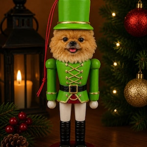 Può includere: Un addobbo a forma di schiaccianoci con la testa di un soffice cane di razza Pomerania marrone che indossa un cilindro verde brillante. La figura indossa una giacca verde con finiture dorate, una cintura nera con fibbia dorata, pantaloni bianchi e stivali neri su una base rossa.