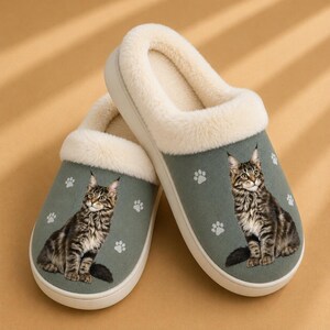 May include: A pair of sage green slip-on slippers featuring a soft cream-colored faux fur lining and collar. The exterior of each slipper is decorated with a printed image of a brown tabby cat and several small white paw print icons.