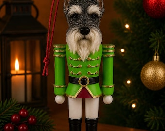 May include: A nutcracker ornament featuring a Schnauzer dog head with grey and white fur. The figure wears a bright green uniform with gold trim, a black belt, white trousers, and black boots, standing on a round red base with a red hanging cord.