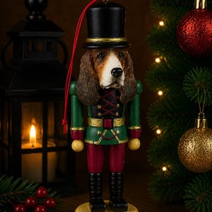 May include: A nutcracker ornament featuring a beagle dog head wearing a black top hat. The figure wears a green jacket with gold trim, red trousers, and black boots, standing on a round yellow base with a red hanging string attached to the top.