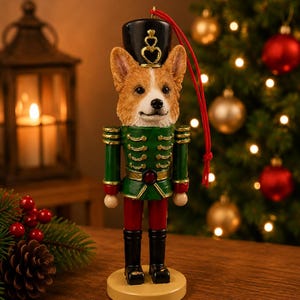 May include: A nutcracker ornament featuring a Corgi dog head with a black hat, a green jacket with gold trim, red trousers, and black boots, standing on a round base with a red hanging cord attached to the top of the hat.