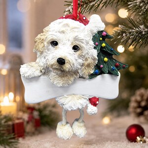 Op de afbeelding: Een hangende kerstversiering in de vorm van een pluizige witte hond met beige oren, die een rode muts met witte stippen draagt. De hond houdt een leeg wit bord in de vorm van een bot vast naast een groene kerstboom met kleurrijke ballen.