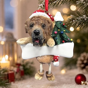 Op de afbeelding: Een kerstboomornament met een bruine hond die een rode kerstmuts met witte rand draagt. De hond staat naast een groene kerstboom met kleurrijke versieringen en houdt een groot, leeg, wit botvormig bordje vast voor persoonlijke tekst.