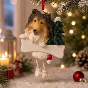 Op de afbeelding: Een hangende kerstversiering met een bruine, zwarte en witte Shetland Sheepdog met een rode kerstmuts. De hond houdt een leeg, wit, botvormig bord vast naast een groene dennenboom. Twee witte pootjes bungelen onder het bord.