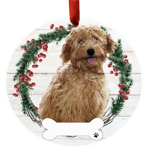 May include: A round Christmas ornament featuring a golden doodle dog sitting in a wreath of green pine branches with red berries. The dog is smiling with its tongue sticking out. The ornament has a white bone with a paw print at the bottom.