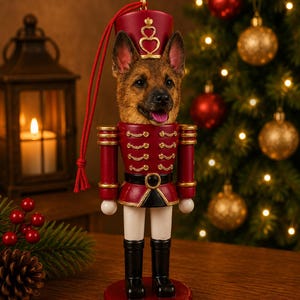 Op de afbeelding: Een notenkraker-ornament met een Duitse herdershondkop op een rood en goud uniform met een witte broek en zwarte laarzen. Het figuur draagt een rode muts met een gouden hartembleem en heeft een rood koord om het ornament op te hangen.