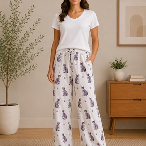 May include: A white V-neck short-sleeved t-shirt paired with white wide-leg pajama pants featuring a repeating pattern of grey cats and small black paw prints. The pants have an elastic waistband with a drawstring tie and side pockets.