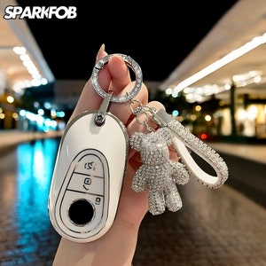 Peut inclure: Un couvre-clé de voiture blanc avec une bordure argentée et trois boutons. Le couvre-clé est attaché à un porte-clés argenté avec un charme en forme d'ours en peluche recouvert de strass.