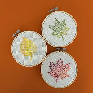 May include: Three embroidery hoops with leaf designs. One features a yellow leaf, another a green leaf, and the third a red and orange leaf. The leaves are stitched on white fabric and set in wooden hoops, perfect for fall decor.