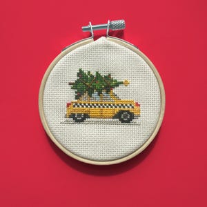 Puede incluir: Bordado de punto de cruz en un aro de madera que representa un taxi amarillo que transporta un árbol de Navidad en su techo. El árbol es verde con detalles rojos y dorados. El taxi tiene un patrón de cuadros blancos y negros.