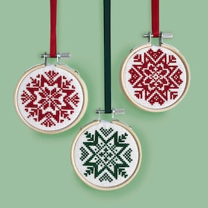 May include: Three round wooden ornaments with red and green snowflake designs embroidered on white fabric. The ornaments are hanging from red, green, and white ribbons.