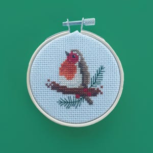 Peut inclure: Broderie au point de croix d'un rouge-gorge sur une branche avec des aiguilles vertes, dans un cercle en bois. L'oiseau a une poitrine rouge et des ailes marron, sur fond bleu clair. Le cercle est rond.
