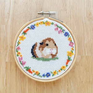 Puede incluir: Un aro de bordado blanco con un conejillo de indias marrón y blanco rodeado de un borde floral colorido.