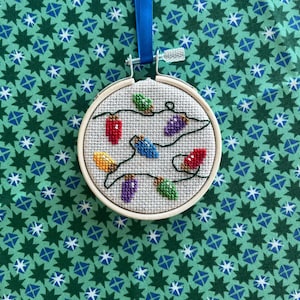 May include: A white circular embroidery hoop with a cross-stitch design of a string of colorful Christmas lights. The hoop is hanging from a blue ribbon against a green background with a white star pattern.