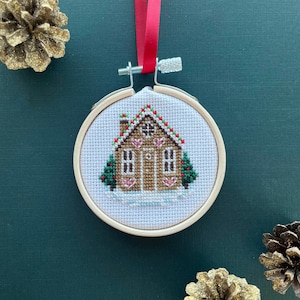 May include: A white, wooden-framed embroidery hoop with a cross-stitch design of a gingerbread house with red and green accents. The house has a brown roof, white trim, and a white door. There are two small green trees on either side of the house.