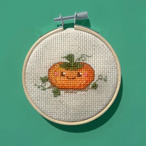 May include: A cross-stitch embroidery of a smiling orange pumpkin with green leaves and vines, set in a wooden embroidery hoop. The pumpkin has rosy cheeks and a happy expression. The background is a light beige fabric.