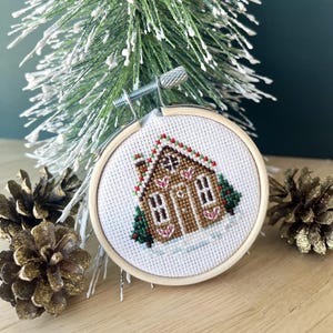 May include: A white cross-stitch embroidery of a gingerbread house with red and green accents, framed in a wooden hoop. The house is decorated with candy canes and hearts.