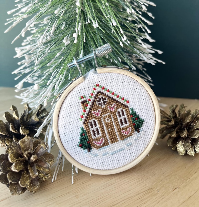 May include: A white cross-stitch embroidery of a gingerbread house with red and green accents, framed in a wooden hoop. The house is decorated with candy canes and hearts.