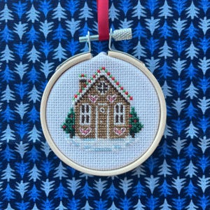 May include: A white cross-stitch ornament featuring a gingerbread house with red and green accents. The house has a brown door and white icing details. The ornament is framed in a wooden hoop.