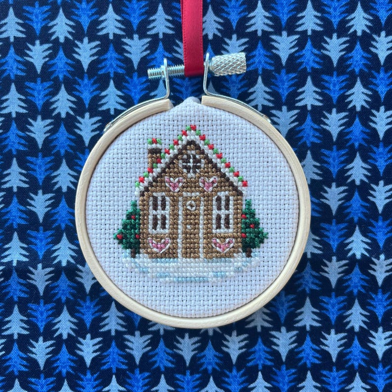 May include: A white cross-stitch ornament featuring a gingerbread house with red and green accents. The house has a brown door and white icing details. The ornament is framed in a wooden hoop.