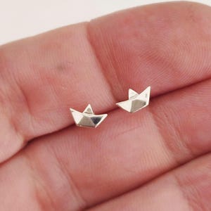 May include: A pair of small, polished silver stud earrings shaped like miniature origami paper boats. Each earring features a pointed bow and stern with a central triangular sail, reflecting light with a smooth, metallic, and reflective surface.