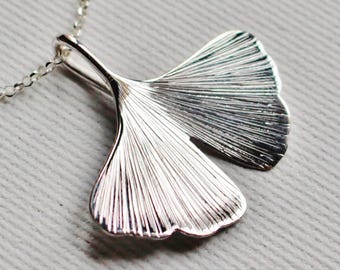 May include: A silver-toned necklace featuring a ginkgo leaf pendant. The pendant has a fan-shaped design with fine, radiating etched lines across its surface. It is attached to a delicate, thin silver-toned chain that extends upwards.