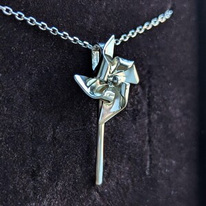 May include: A silver-toned pendant shaped like a pinwheel attached to a delicate link chain. The pinwheel features four curved blades radiating from a central point, with a smooth, reflective metallic finish throughout the entire piece.