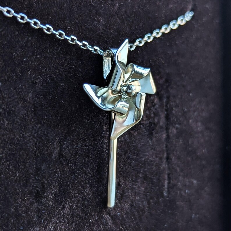 May include: A silver-toned pendant shaped like a pinwheel attached to a delicate link chain. The pinwheel features four curved blades radiating from a central point, with a smooth, reflective metallic finish throughout the entire piece.